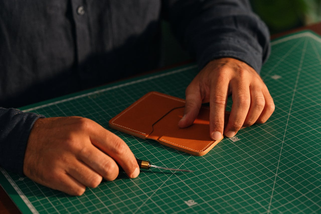 Close-up of hands crafting a leather wallet on a green cutting mat, emphasizing artisanal craftsmanship.